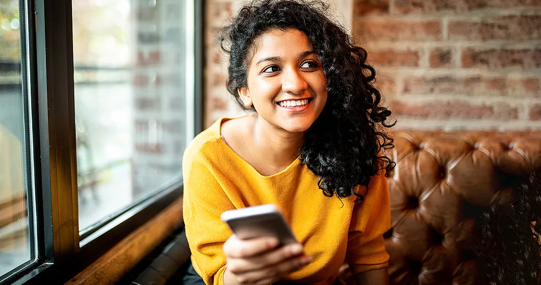 Lächelnde junge Frau sitzt auf einem Sofa und sucht eine Domain auf einem Smartphone